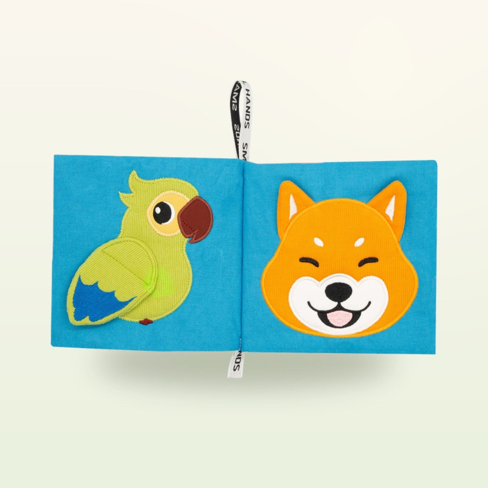 Small Hands Haustiere Montessori Stoffbuch aufgeschlagen – grüner Papagei und lächelnder Shiba-Hund auf sanftem Creme-Hintergrund – ab 0 Monaten