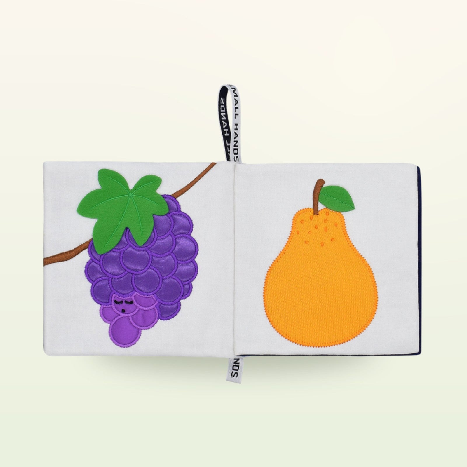 Small Hands Bunte Früchte Montessori Stoffbuch aufgeschlagen – lila Weintraube und orange Birne auf sanftem Creme-Hintergrund – ab 0 Monaten