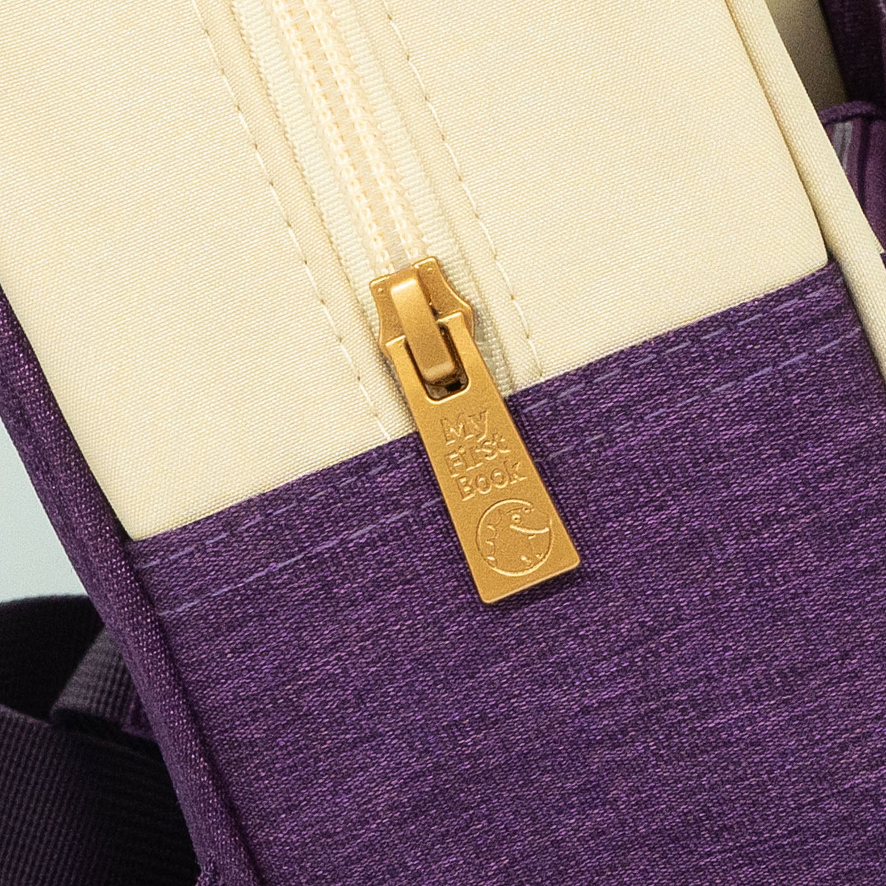 Detailaufnahme des Märchenwelten Einhorn-Rucksacks: goldfarbener Reißverschluss mit eingeprägtem „My First Book“-Logo auf creme-lila Stoff – zeigt die hochwertige Verarbeitung. Ab 1 Jahr.