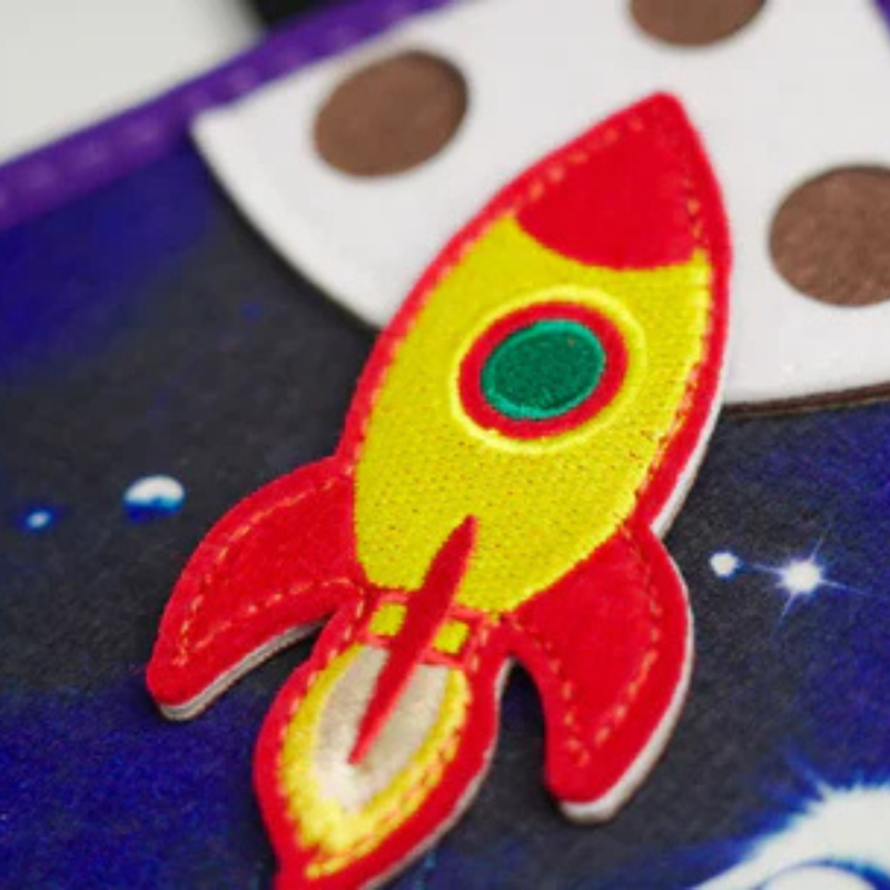 Detailaufnahme der roten Filz-Rakete mit grünem Bullauge auf Sternenhimmel im Weltraum Quiet Book – hochwertige Verarbeitung