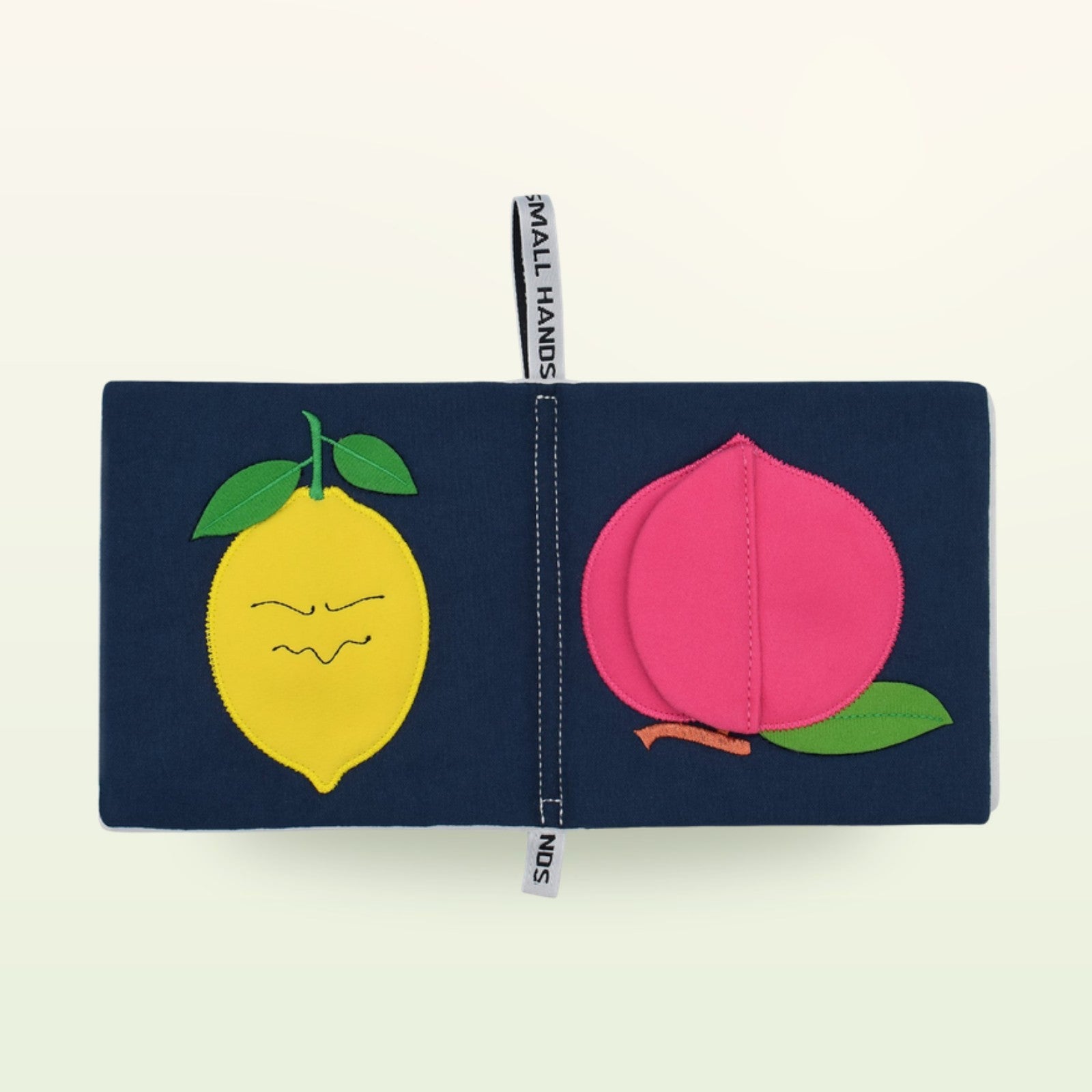 Small Hands Bunte Früchte Montessori Stoffbuch aufgeschlagen – gelbe Zitrone mit Gesicht und pinker Pfirsich auf sanftem Creme-Hintergrund – ab 0 Monaten