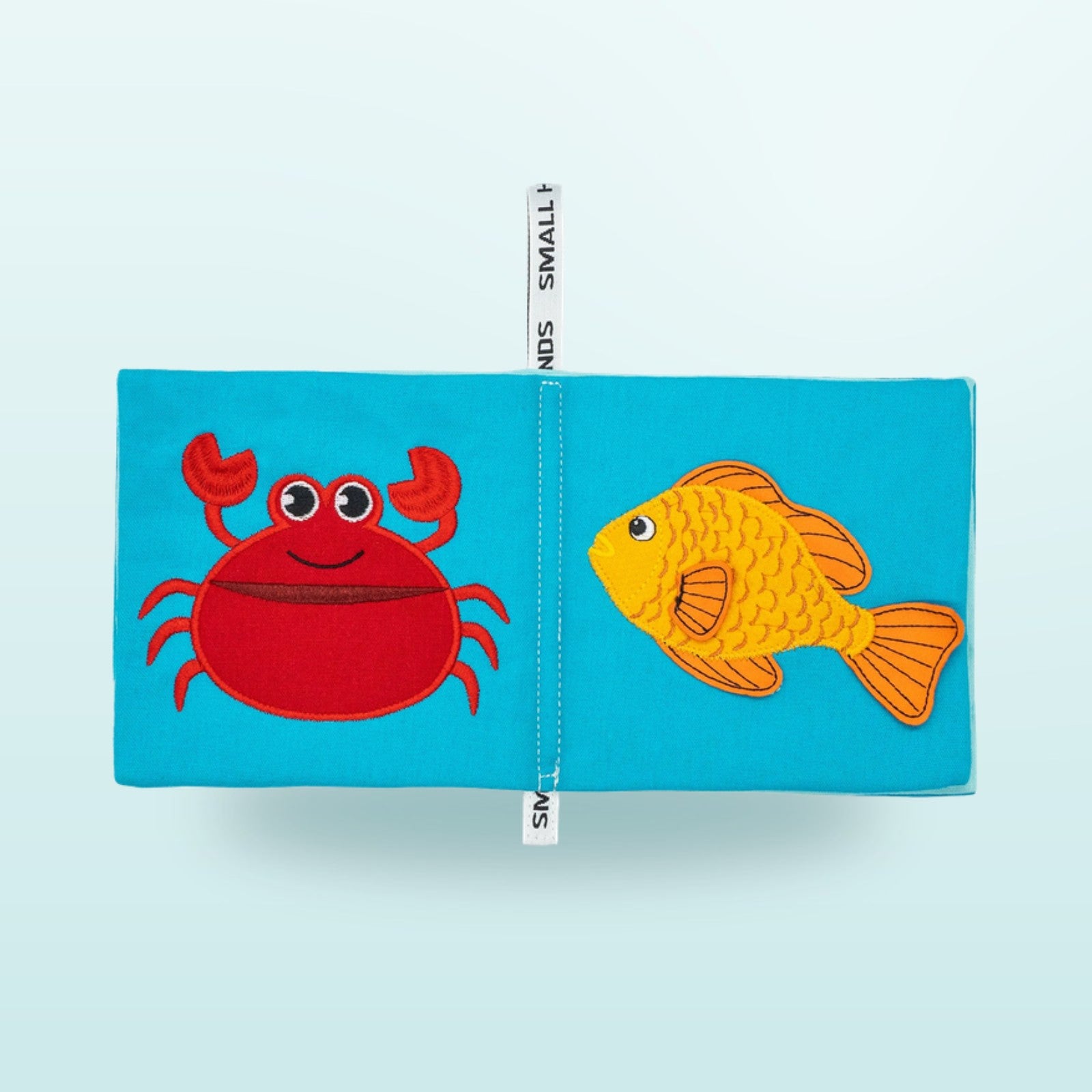 Small Hands Meeresbewohner Montessori Stoffbuch Doppelseite mit Krabbe und Goldfisch auf sanftem Unterwasser-Blau-Hintergrund – ab 1 Jahr
