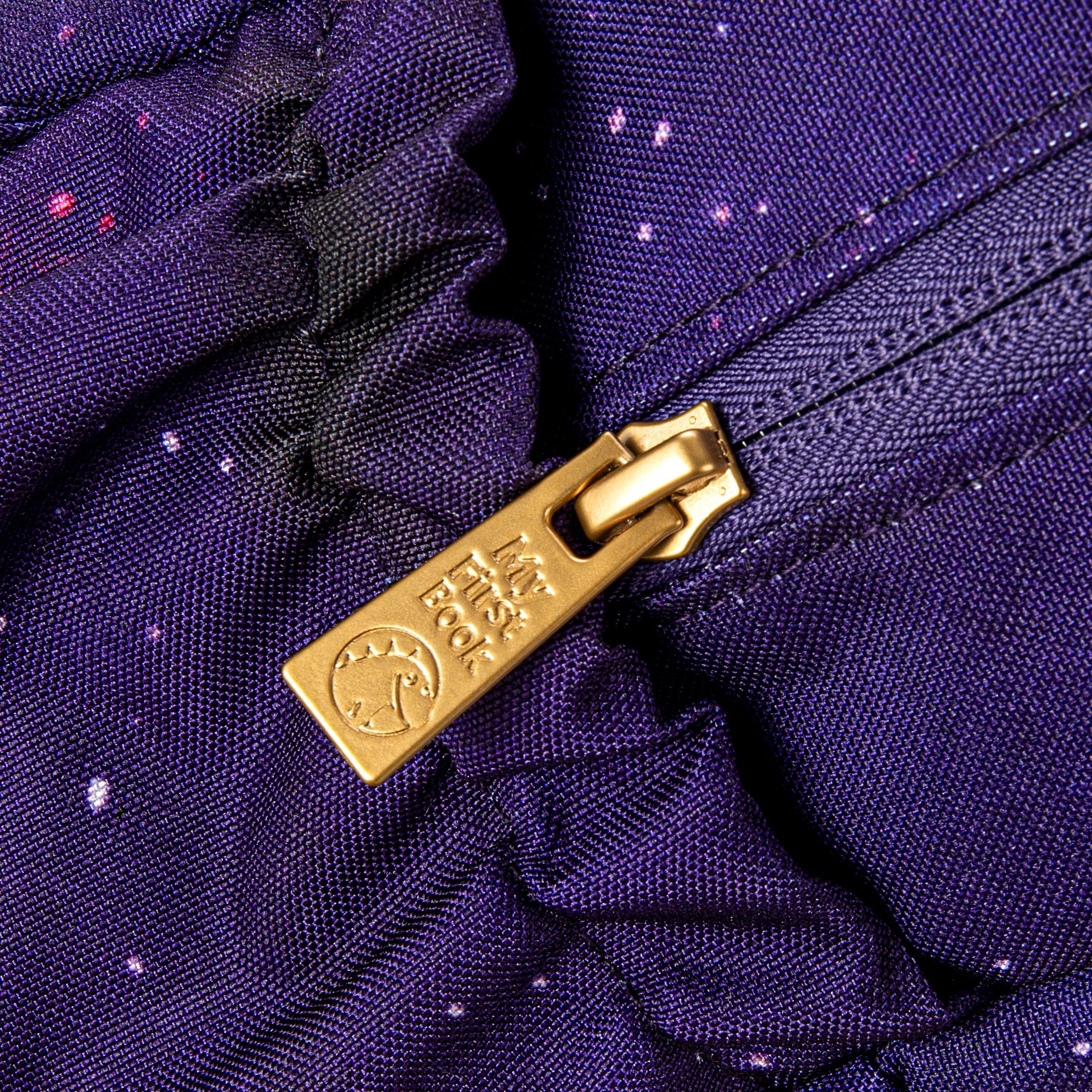 Detailaufnahme des goldenen Reißverschlusses mit -My First Book- Logo am lila Galaxie-Weltraum-Rucksack