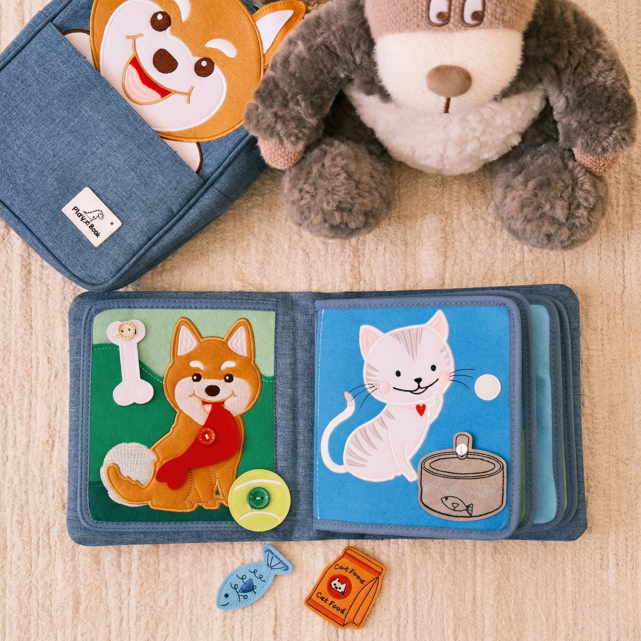 Aufgeschlagenes Pet Friends Quiet Book: links Hund mit Knochen, rotem Knopf-Halstuch und Tennisball, rechts Katze mit Napf und Fisch; im Hintergrund Plüschhund und Shiba-Rucksack, davor lose Filz-Teile. Ab 1 Jahr.